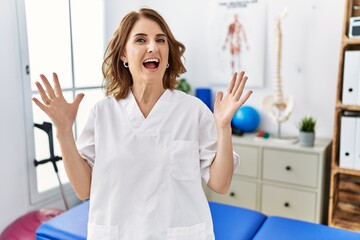 Middle age physiotherapist woman working at pain recovery clinic celebrating surprised and amazed for success with arms raised and open eyes. winner concept.