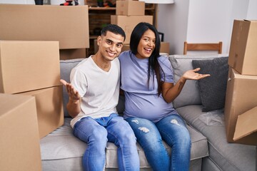 Young hispanic couple expecting a baby sitting on the sofa at new home celebrating achievement with happy smile and winner expression with raised hand