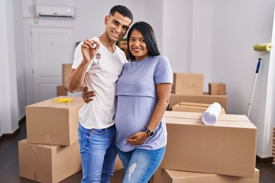 Young latin couple expecting baby hugging each other holding key at new home