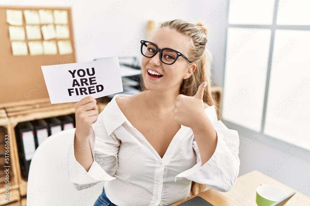 Sticker young caucasian woman holding you are fired banner at the office smiling happy and positive, thumb u