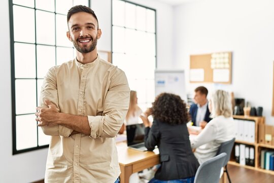 Young hispanic businessman smiling happy standing with arms crossed gesture at the office during business meeting.