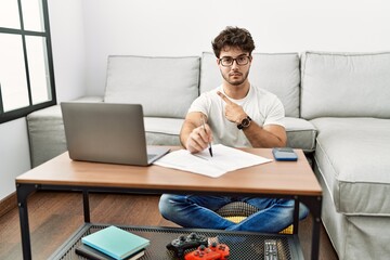 Fototapeta premium Hispanic man doing papers at home pointing with hand finger to the side showing advertisement, serious and calm face