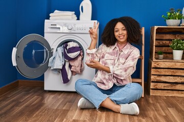 Young african american woman sitting on the floor doing laundry smiling with happy face winking at...
