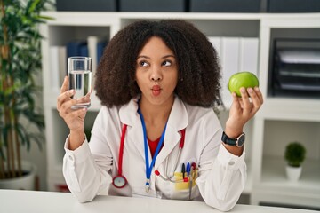 Young african american dietitian woman holding fresh apple and water making fish face with mouth...