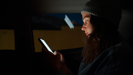 Young attractive woman takes a ride in taxi car at night city, holding cell phone, checking on mobile, using smartphone, scrolling social media news, reading email letters, messages, shopping online