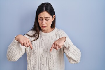 Young brunette woman standing over blue background pointing down looking sad and upset, indicating...