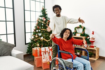 Young interracial couple with woman sitting on wheelchair by christmas tree looking at the camera smiling with open arms for hug. cheerful expression embracing happiness.