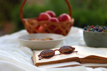 Picninc blanket with straw bag, bowl of strawberries and blueberries, bowl of chocolate chip cookies, books, sunglasses and basket of apples in the garden. Selective focus.