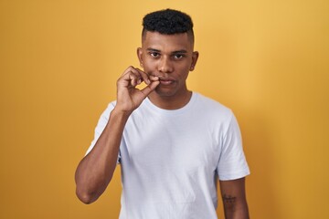 Young hispanic man standing over yellow background mouth and lips shut as zip with fingers. secret...