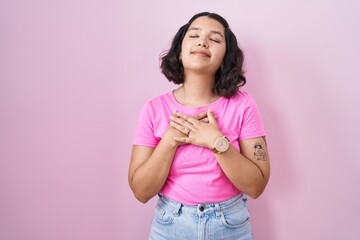 Young hispanic woman standing over pink background smiling with hands on chest with closed eyes and grateful gesture on face. health concept.