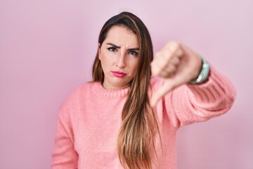 Fototapeta premium Young hispanic woman standing over pink background looking unhappy and angry showing rejection and negative with thumbs down gesture. bad expression.