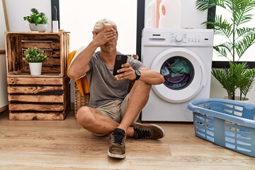Young blond man doing laundry using smartphone covering eyes with hand, looking serious and sad....