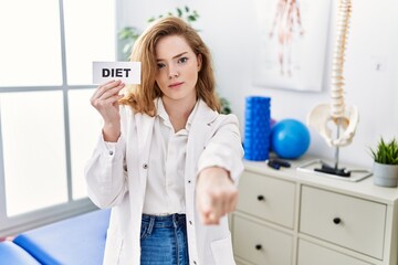 Young caucasian woman working at rehabilitation clinic holding diet banner pointing with finger to the camera and to you, confident gesture looking serious