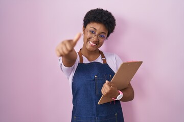 Young african american woman wearing professional waitress apron holding clipboard approving doing...