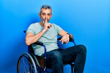 Fototapeta premium Handsome middle age man with grey hair sitting on wheelchair asking to be quiet with finger on lips. silence and secret concept.