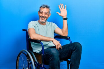 Handsome middle age man with grey hair sitting on wheelchair waiving saying hello happy and...