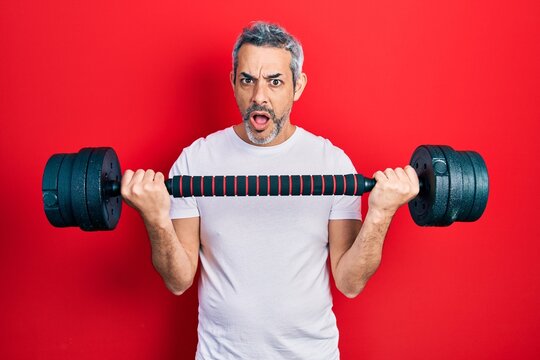 Handsome middle age man with grey hair wearing sportswear using dumbbells in shock face, looking skeptical and sarcastic, surprised with open mouth