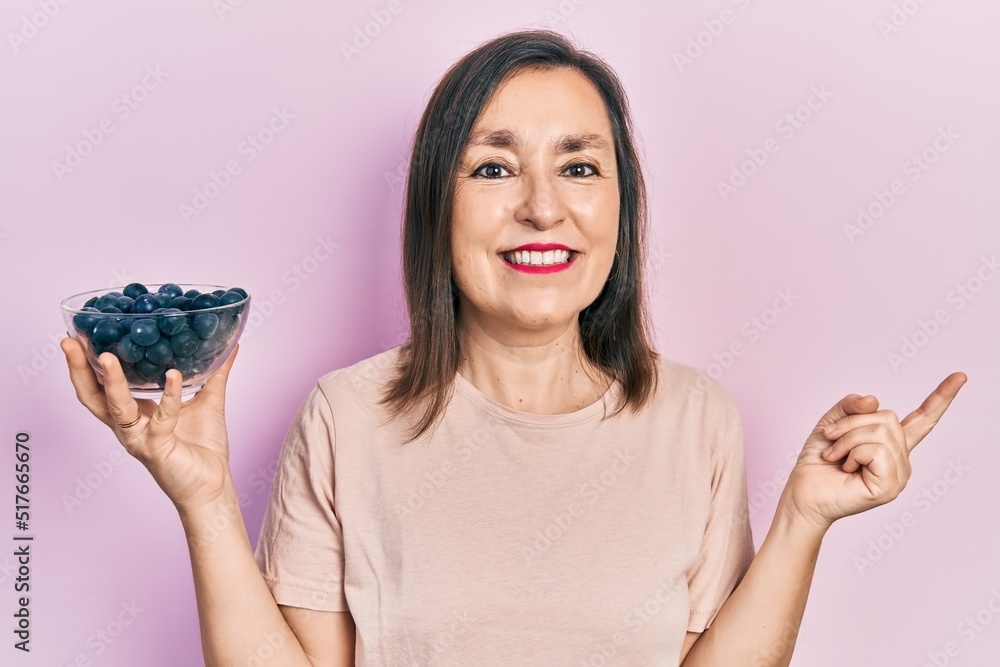 Wall mural middle age hispanic woman holding blueberries smiling happy pointing with hand and finger to the sid