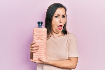 Middle age hispanic woman holding detergent bottle in shock face, looking skeptical and sarcastic, surprised with open mouth