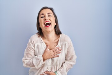 Middle age hispanic woman standing over blue background smiling and laughing hard out loud because funny crazy joke with hands on body.