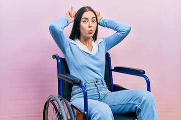 Beautiful woman with blue eyes sitting on wheelchair doing funny gesture with finger over head as...