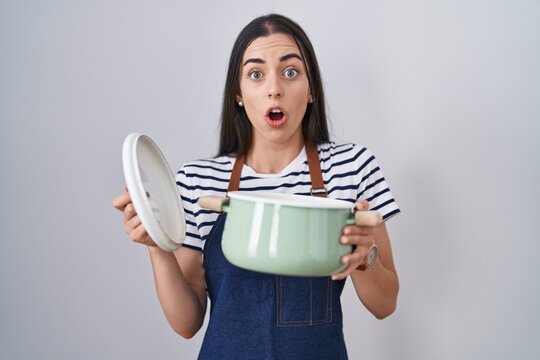 Young brunette woman wearing apron holding cooking pot afraid and shocked with surprise and amazed expression, fear and excited face.