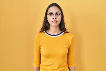 Obraz premium Young brazilian woman wearing glasses over yellow background with serious expression on face. simple and natural looking at the camera.