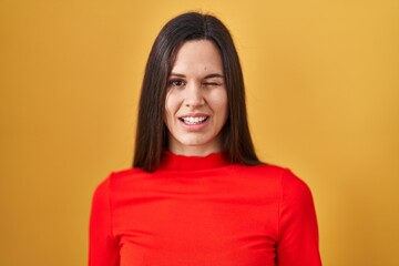Young hispanic woman standing over yellow background winking looking at the camera with sexy...