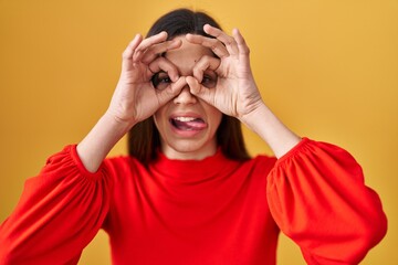 Young hispanic woman standing over yellow background doing ok gesture like binoculars sticking tongue out, eyes looking through fingers. crazy expression.