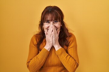 Middle age hispanic woman standing over yellow background laughing and embarrassed giggle covering...