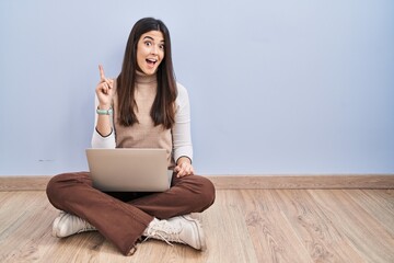Young brunette woman working using computer laptop sitting on the floor pointing finger up with successful idea. exited and happy. number one.