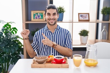 Fototapeta na wymiar Hispanic man with long hair sitting on the table having breakfast smiling and looking at the camera pointing with two hands and fingers to the side.