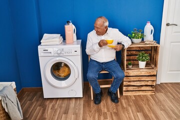 Senior man drinking coffee waiting for washing machine at laundry room