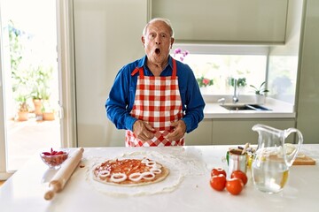 Senior man with grey hair cooking pizza at home kitchen scared and amazed with open mouth for surprise, disbelief face