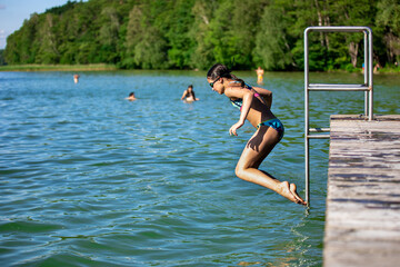 Preteen sporty girl jumping into water of lake from wooden pier on hot sunny summer da