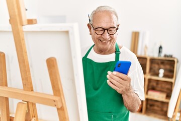 Senior grey-haired artist man smiling happy and using smartphone painting at art studio.