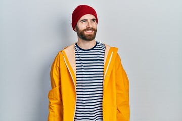 Caucasian man with beard wearing yellow raincoat looking away to side with smile on face, natural expression. laughing confident.