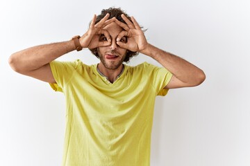 Young hispanic man standing over isolated background doing ok gesture like binoculars sticking tongue out, eyes looking through fingers. crazy expression.