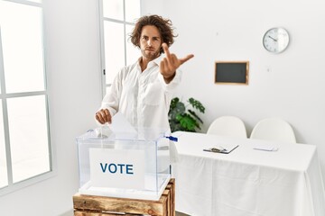 Young hispanic man voting putting envelop in ballot box showing middle finger, impolite and rude...