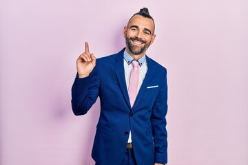 Young hispanic man wearing business suit and tie pointing finger up with successful idea. exited and happy. number one.