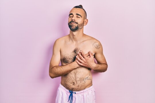 Young hispanic man shirtless wearing swimsuit smiling with hands on chest with closed eyes and grateful gesture on face. health concept.