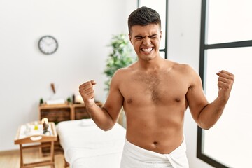 Obraz premium Young hispanic man standing shirtless at spa center very happy and excited doing winner gesture with arms raised, smiling and screaming for success. celebration concept.