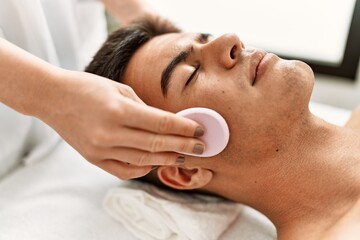 Young hispanic man relaxed having facial treatment cleaning face with sponge at beauty center
