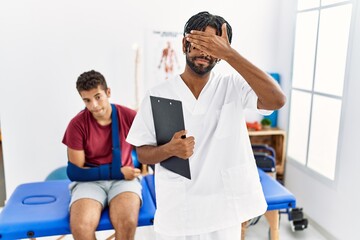 Young hispanic man working at pain recovery clinic with a man with broken arm covering eyes with...