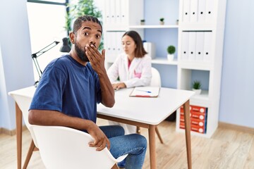 Obraz premium Young hispanic man at doctor clinic covering mouth with hand, shocked and afraid for mistake. surprised expression