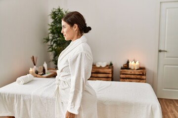 Young beautiful hispanic woman wearing bathrobe at wellness spa looking to side, relax profile pose with natural face with confident smile. - obrazy, fototapety, plakaty