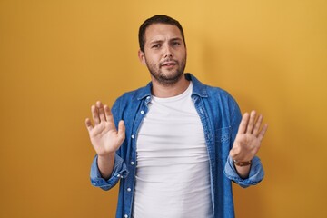 Hispanic man standing over yellow background moving away hands palms showing refusal and denial...