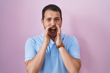 Hispanic man standing over pink background shouting angry out loud with hands over mouth