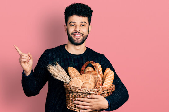Young arab man with beard holding wicker basket with bread smiling happy pointing with hand and finger to the side
