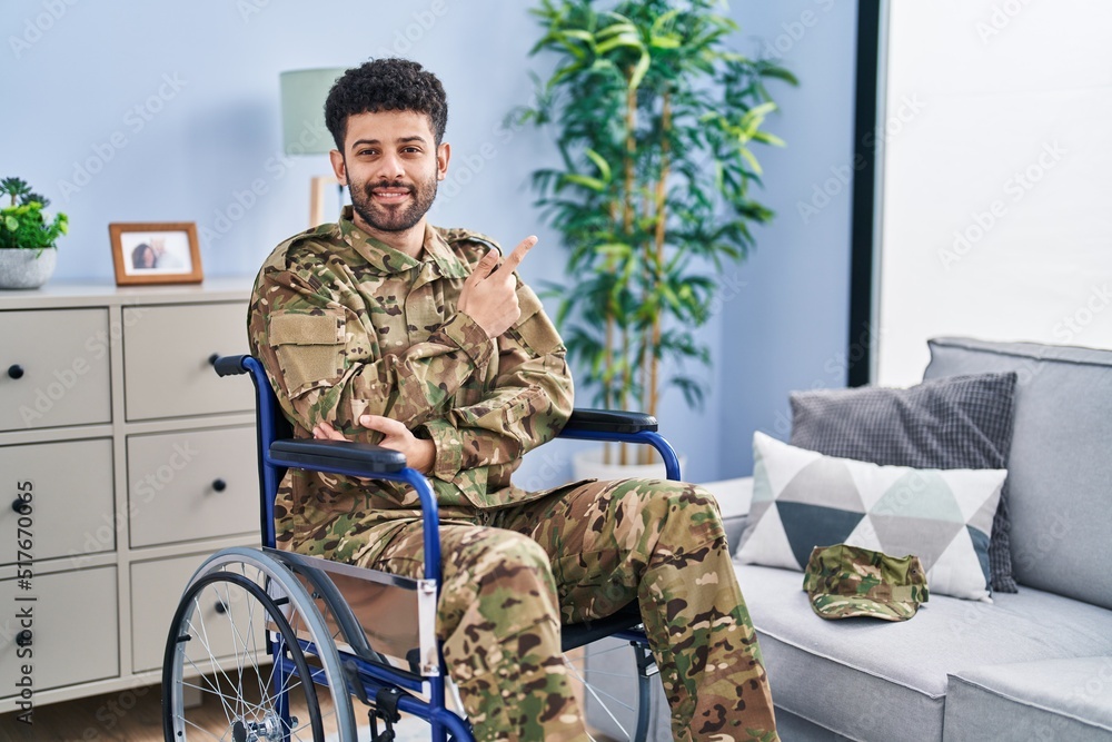 Poster Arab man wearing camouflage army uniform sitting on wheelchair with a big smile on face, pointing with hand and finger to the side looking at the camera.